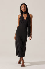 A woman is wearing a sleeveless, black, V-neck evening dress with gathered fabric detailing around the neckline. The dress is fitted at the waist and flows down to a midi length. She has paired the dress with a matching black scarf draped over her neck and black strappy high-heeled sandals.