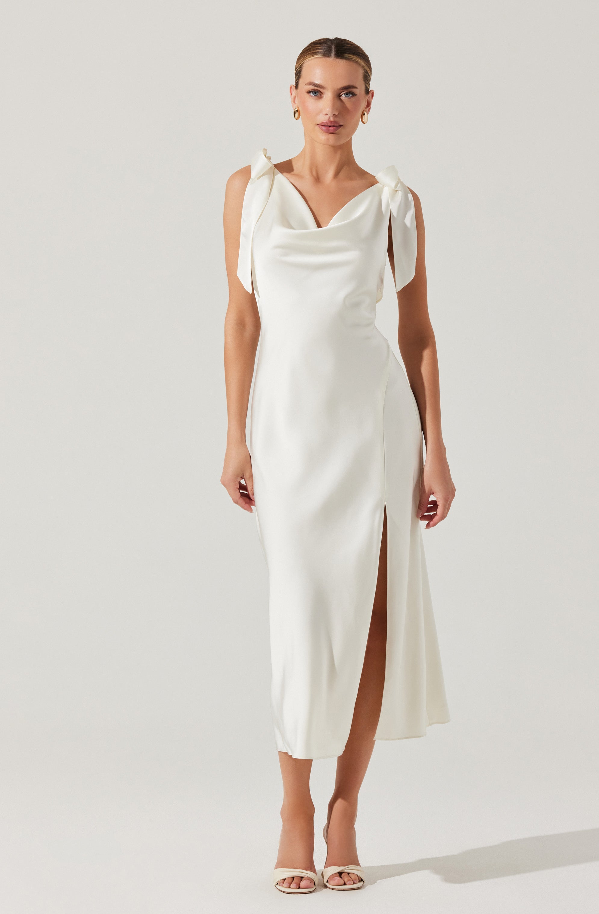 A woman is wearing a long, white, elegant dress with shoulder straps tied into bows. The dress has a cowl neckline and a side slit that reveals her leg. She is also wearing open-toed, high-heeled sandals and gold hoop earrings.