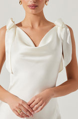 The dress is made of a smooth, ivory satin fabric with a subtle sheen. It features a sleeveless design with shoulder details that are tied into bows, creating a soft, feminine look. The neckline is draped in a cowl style, adding elegance to the overall design.