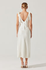 The woman is wearing a sleeveless, white, ankle-length dress with a deep V neckline at the back. The dress features delicate shoulder straps tied into small bows. She is also wearing light-colored high-heeled shoes.