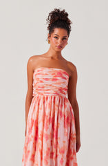 A woman is wearing a strapless dress with a fitted, ruched bodice and a flowing skirt. The fabric features a pink and orange tie-dye pattern, creating a soft, watercolor effect.