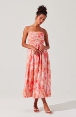 A woman is wearing a strapless maxi dress with a fitted bodice featuring horizontal ruched stripes in shades of pink and orange. The skirt is flowy, with a watercolor-like pattern of pink and orange tie-dye in a midi length. She is also wearing open-toe white sandals with thin straps.