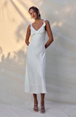A woman is wearing a long, white satin dress with ruffled shoulder straps and a ruffled neckline. The dress is fitted at the waist and flows down to her ankles with a slight flare at the hem. She is also wearing white strappy high-heeled sandals.