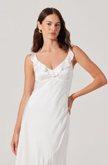 A woman is wearing a sleeveless, white satin dress with ruffled details along the V-neckline. The dress is smooth and shiny, with a relaxed fit that drapes elegantly. She accessorizes with gold earrings and has shoulder-length brown hair styled in soft waves.
