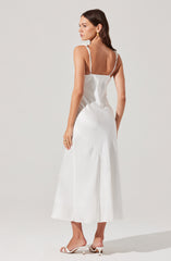 A woman is wearing a white, sleeveless, ankle-length dress with thin, adjustable straps. The dress has a fitted bodice and a slightly flared skirt with a gathered detail around the hips. She is also wearing white heeled sandals and gold earrings.