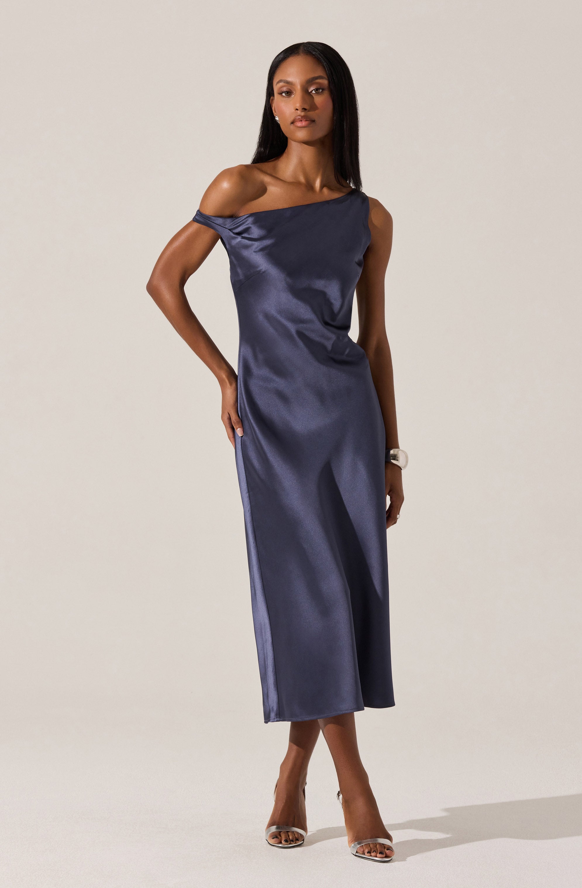 A woman is wearing a sleeveless, off-the-shoulder navy blue satin dress that falls to mid-calf length. The fabric has a smooth, shiny finish, and the dress features a loose, relaxed fit. She completes the look with clear high-heeled shoes and a wristwatch.