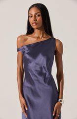 A woman is wearing a navy blue satin dress with an asymmetrical neckline that drapes over one shoulder, leaving the other shoulder bare. The fabric has a smooth, shiny texture that reflects light. She accessorizes with a chunky silver bracelet on her right wrist.
