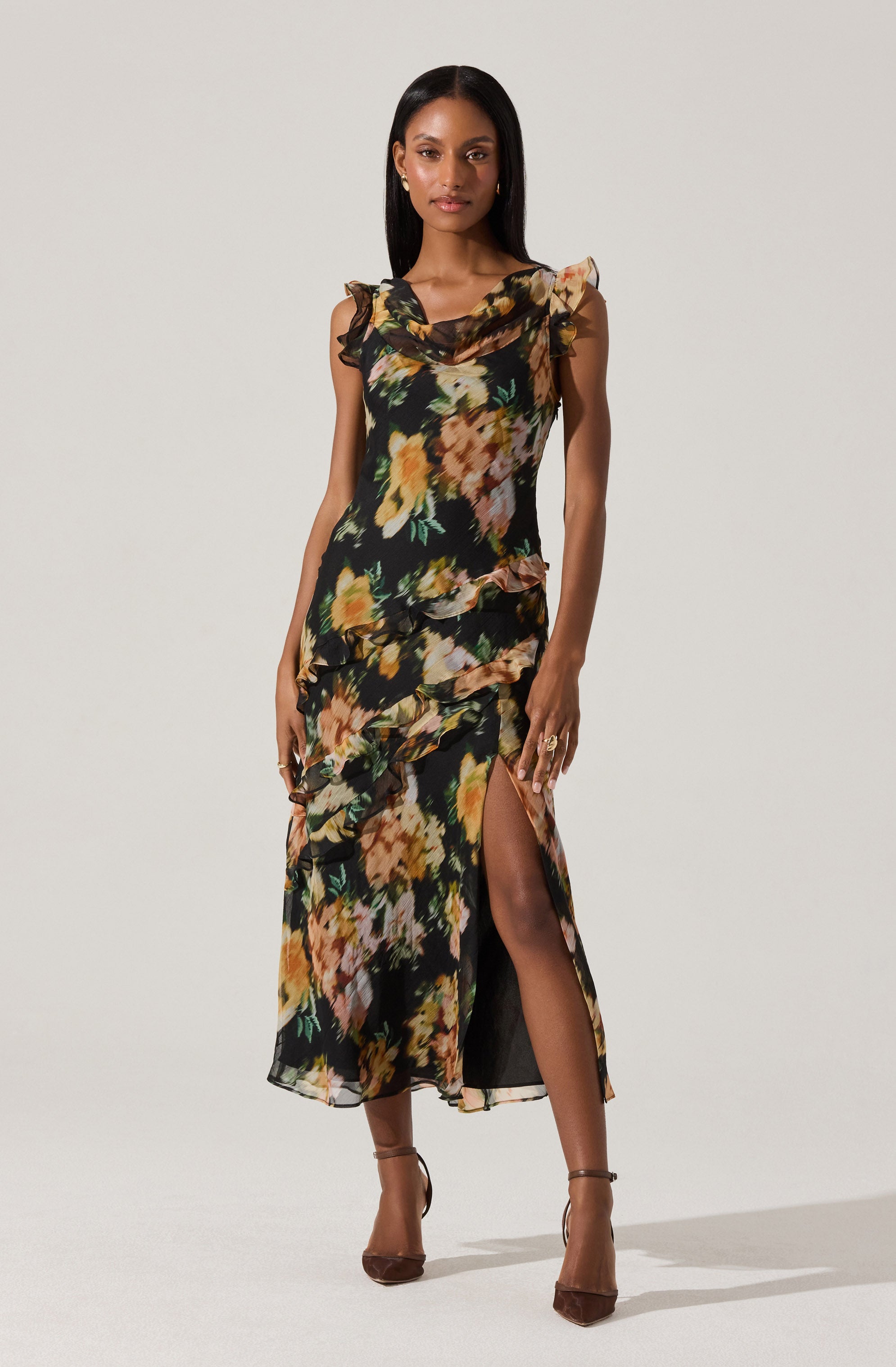 A woman is wearing a sleeveless, floor-length dress with a black background and a large, multicolored floral pattern. The dress features ruffled details around the shoulders and edges, and has a high slit on one side revealing her leg. She is wearing high-heeled shoes and has minimal accessories.