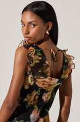 A woman is wearing a sleeveless dress with ruffled shoulder straps tied at the back with thin strings. The dress has a dark background with a multicolored, abstract floral pattern featuring shades of orange, green, and beige. She is accessorized with large, gold, oval-shaped earrings, and her hair is sleek and straight.