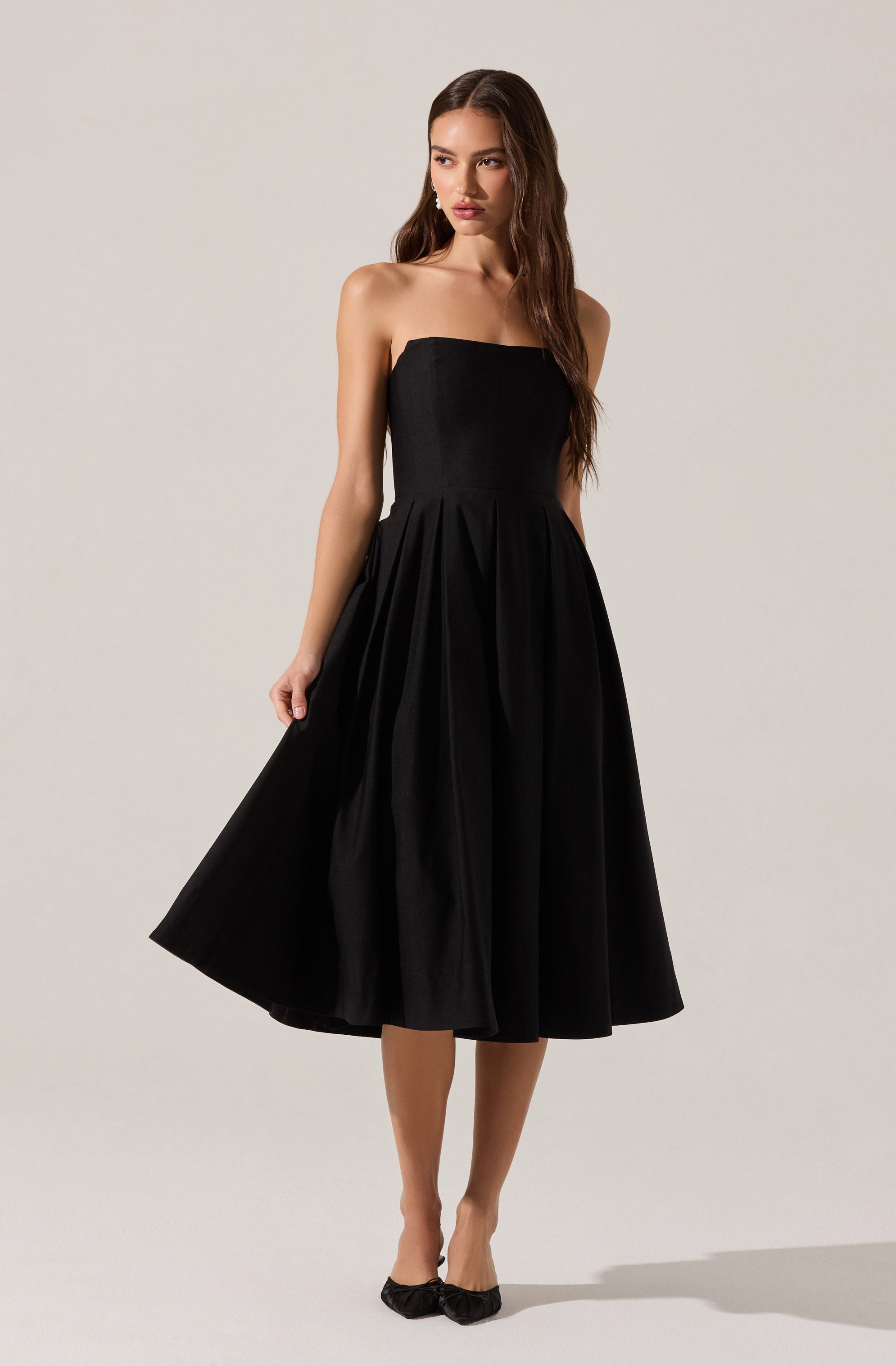 A woman is wearing a strapless, black midi dress with a fitted bodice and a full, pleated skirt that falls below the knees. She is also wearing black shoes with decorative bows at the front.