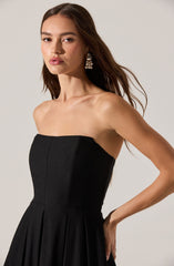 A woman is wearing a strapless black dress with a fitted bodice and a pleated skirt. She has long brown hair and is accessorized with large, dangling pearl earrings.