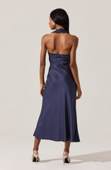 A woman is wearing a sleeveless, halter-neck satin dress in a deep navy blue color that extends to her ankles. The dress features a fitted bodice with slight ruching at the waist and a smooth, flowing skirt. She is also wearing high-heeled, open-toe sandals with ankle straps.