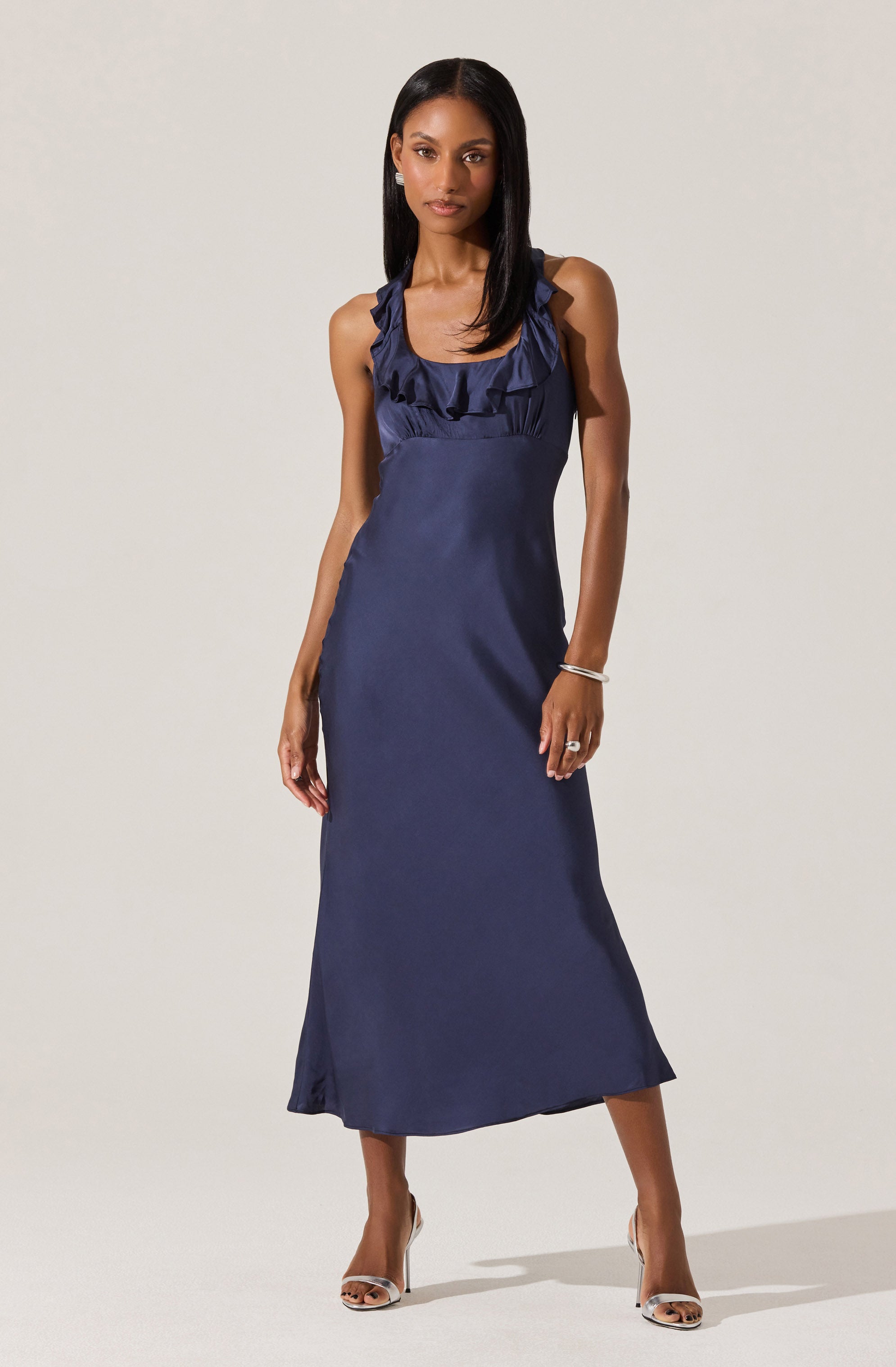 A woman is wearing a sleeveless, navy blue, satin evening dress with ruffled detailing around the neckline and shoulders. The dress is fitted at the waist and flows down to a slightly flared hemline reaching mid-calf. She is accessorizing with simple jewelry and open-toed heels.