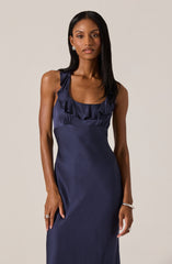 A woman is wearing a sleeveless, navy blue satin dress with a ruffled neckline. The dress has a fitted waist and a smooth, shiny fabric that extends down. She accessorizes with earrings, rings, and a bracelet.