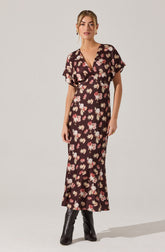 A woman is wearing a long, dark-colored dress with a floral pattern featuring pink, white, and beige flowers. The dress has a V-neckline, short loose sleeves, and a fitted waist. It extends down to her ankles and she is wearing black ankle boots underneath.