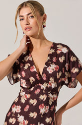 A woman is wearing a dress with a deep V-neckline and short, loose sleeves. The dress features a dark background with a floral pattern in shades of pink, cream, and beige.