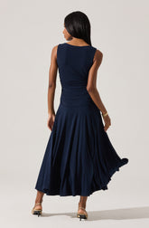 A woman is wearing a sleeveless, navy blue dress with a fitted bodice that gathers at the waist. The skirt is long and flowing with pleats, creating a full, elegant silhouette. She has accessorized with a gold bracelet and is wearing high-heeled shoes.