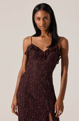 A woman is wearing a sleeveless, form-fitting dress made of burgundy lace fabric. The dress features thin spaghetti straps and ruffled detailing along the neckline. The lace fabric has a floral pattern, and the dress has a front slit at the bottom.