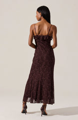A woman is wearing a long, burgundy lace dress with thin spaghetti straps. The dress features a ruffle detail along the top edge and has a fitted silhouette that flares slightly at the hem. She is also wearing dark, open-toe high-heeled shoes.