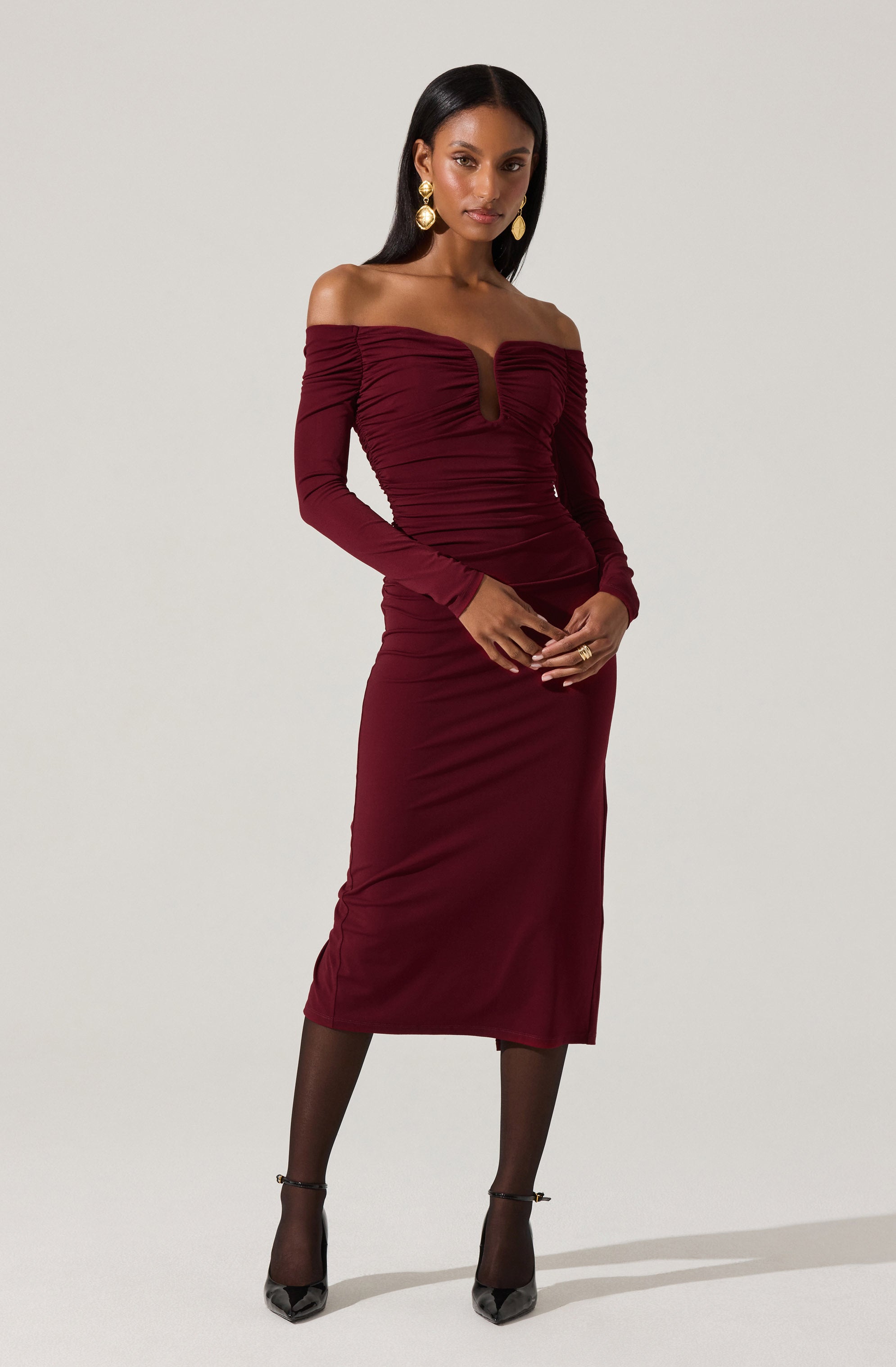 A woman is wearing an elegant, off-the-shoulder, long-sleeved dress in a deep burgundy color. The dress features a ruched design across the bodice and skirt, with a keyhole cutout at the center of the neckline. She accessorizes with large, gold drop earrings and black sheer stockings paired with black pointed-toe heels.