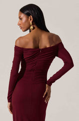 A woman is wearing an off-the-shoulder, long-sleeved maroon gown with ruched detailing along the bodice. The dress is fitted and extends down to the floor. She accessorizes with large, gold, geometric-shaped earrings and has long, straight black hair.