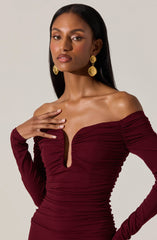 A woman is wearing an off-the-shoulder, long-sleeved dress in a deep burgundy color. The dress features ruching details across the torso and has a plunging neckline. She accessorizes with large, gold statement earrings.