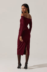 A woman is wearing a fitted, off-the-shoulder, long-sleeve burgundy dress with ruched details along the sides and sleeves. The dress has a front slit at the hem, revealing her black sheer tights and high-heeled shoes.