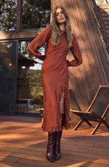A woman is wearing a long, rust-colored dress with sheer, puffed sleeves and lace trim details. The dress features a slit down the front, revealing tall, dark brown knee-high boots.