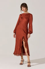 A woman is wearing a long, rust-colored satin dress with a subtle sheen. The dress features long, puffed sleeves gathered at the wrists, a fitted bodice, and a high neckline. It has a front slit that extends from the hem to mid-thigh, adorned with delicate lace trim along the edges of the slit and hemline. The dress has a smooth texture with a slight crinkled effect and appears to be designed for an elegant occasion. She completes her outfit with open-toe high-heeled sandals with ankle straps tied in bows.