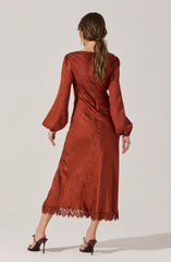 A woman is wearing a long, rust-colored satin dress with a subtle floral pattern. The dress features long, puffed sleeves with elasticized cuffs and a scalloped lace hem at the bottom.