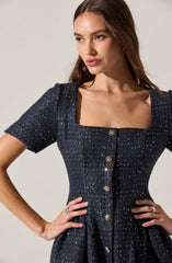 A woman is wearing a dark blue, textured dress with short sleeves. The dress has a square neckline and features a row of decorative gold buttons down the front. The fabric of the dress has a sparkly, glittery appearance.