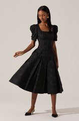 A woman wearing a black midi dress with puffed short sleeves and a square neckline. The dress features a textured fabric and pleats that add volume and movement. She is also wearing black shoes with a bow detail on the toes.