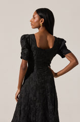 A woman is wearing a black textured dress with short, puffed sleeves and a square neckline. The fabric of the dress appears to have a subtle sheen and a crinkled or embossed pattern. She accessorized with large, round gold earrings.