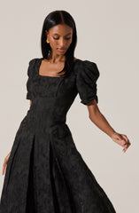 A woman is wearing a black dress with a square neckline, puffed short sleeves, and a textured fabric with a subtle floral pattern. The dress features a fitted bodice and pleats that run down the front of the skirt, creating a flowing silhouette. She has gold earrings and dark, straight hair.