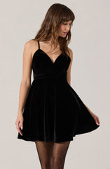 A woman is wearing a sleeveless, black velvet dress with thin spaghetti straps. The dress has a fitted bodice with a V-neckline and a flared skirt that falls above the knees. She is also wearing sheer black tights.