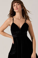A woman is wearing a black, sleeveless dress with thin spaghetti straps. The dress features a V-neckline and a fitted bodice, made from a shimmering, velvety fabric. The skirt portion of the dress flows down smoothly, creating an elegant silhouette.