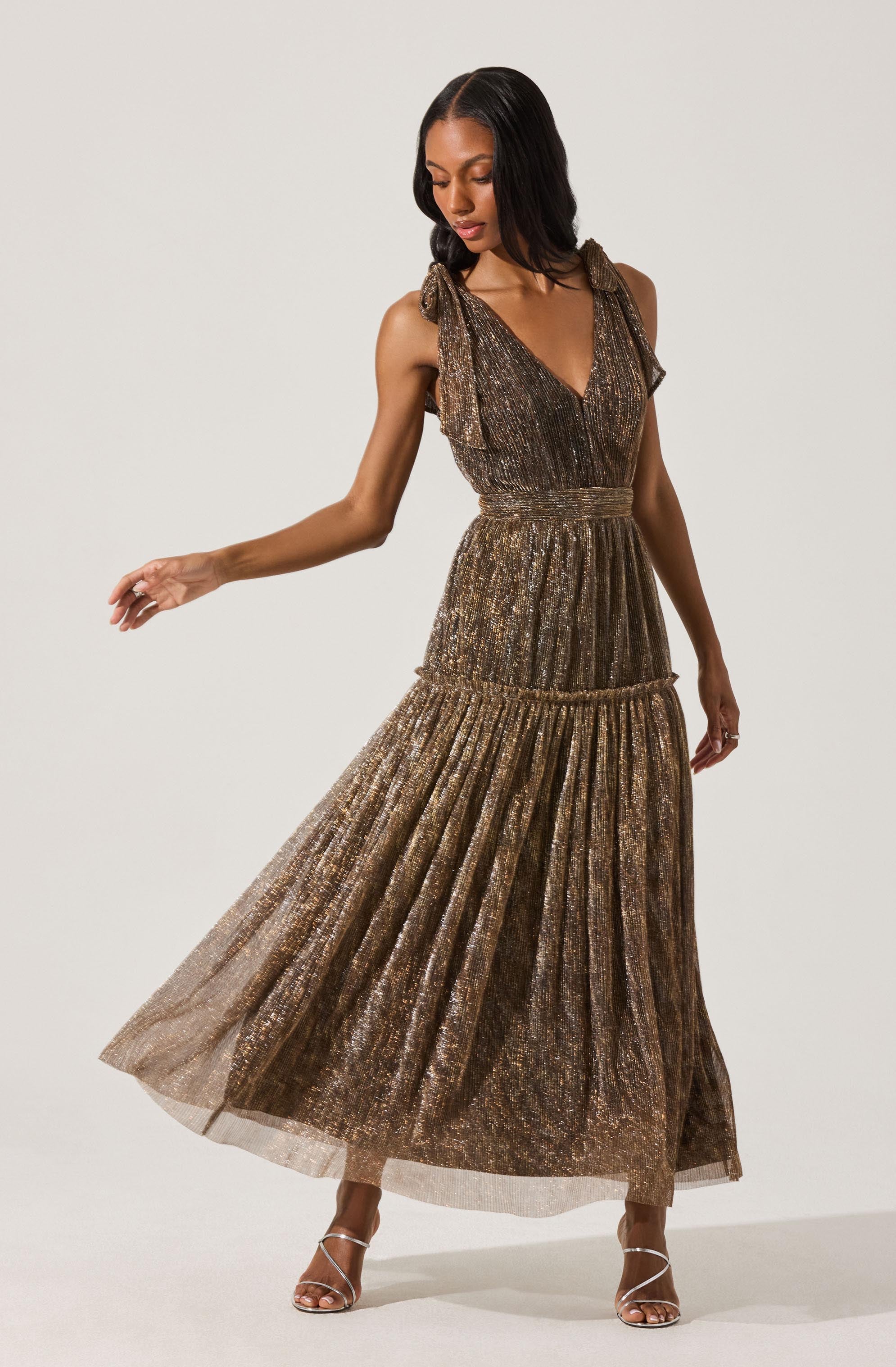 A woman is wearing a sleeveless, metallic, gold and bronze dress with a pleated tulle skirt. The dress features fabric shoulder ties and a fitted bodice that transitions into a flowing, ruffled skirt. She is also wearing strappy high-heeled heels.