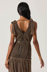 The dress features a sleeveless design with shoulder ties and a deep V-back. It is made of a shimmery, metallic fabric with vertical stripes in gold, bronze, and black tones. The waist is cinched with a matching fabric belt, and the skirt has a layered, ruffled detail towards the bottom.