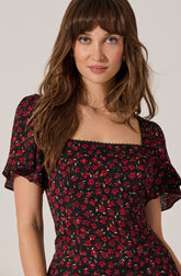 A woman is wearing a black dress with a floral pattern of small red flowers and green leaves. The dress has ruffled, semi-sheer short sleeves and a square neckline with a lace trim.