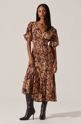 A woman is wearing a long, flowy dress with a leopard print pattern in shades of tan and black. The dress has puffed short sleeves and a V-neckline. She has paired the dress with tall black heeled boots and is accessorizing with a gold bracelet.