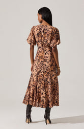 A woman wearing a long, flowy dress with a leopard print pattern in shades of brown and black. The dress features puffed short sleeves and a fitted waist. She is also wearing high-heeled black boots and gold jewelry.