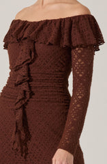 A woman is wearing an elegant off-the-shoulder dress made of brown lace fabric. The dress features a large ruffle that runs along the neckline and extends around the shoulders, adding a decorative touch. The long sleeves are fitted and made from the same lace material, which has a floral pattern. The dress is form-fitting, with ruching details on the sides.