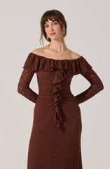 A woman is wearing an off-the-shoulder, long-sleeved dress in a dark brown, lace or textured fabric. The dress features a ruffled neckline with a matching ruffle detail running vertically down the front.
