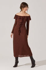 A woman is wearing a long, off-the-shoulder, ruffled dress in a rich brown color. The dress features a textured, lace-like pattern with a ruffle detail that cascades down the front. She pairs the dress with tall black boots.