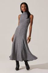 A woman is wearing a sleeveless, high-neck, gray knit dress that is fitted at the top and flows loosely to the floor. She is also wearing black knee-high boots, gold hoop earrings, and gold bracelets on her right wrist.