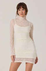 A woman is wearing a fitted, long-sleeve, cream-colored lace dress. The dress features a high neckline and is made of intricate lace fabric with a floral pattern. The dress is semi-sheer, with a lining that covers the torso and extends to just above the knees, creating a textured and elegant look.