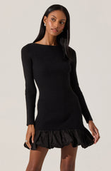 A woman is wearing a fitted, black, long-sleeve, ribbed knit dress with a round neckline. The dress features a ruffled, voluminous hem at the bottom.