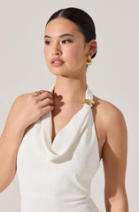 A woman is wearing a sleeveless white top with a draped neckline. She has gold earrings, a gold ring, and a gold bracelet. The accessories have a shiny, metallic finish.