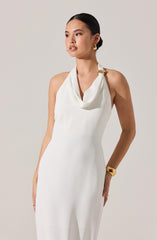 A woman is wearing a sleeveless, white dress with a halter neckline that drapes in a loose, cowl style around her neck, secured with a large, decorative gold ring on the right shoulder. She has minimalist gold jewelry, including a pair of hoop earrings, a wide bracelet on her right wrist, and a thin ring on her left hand.