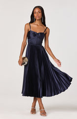 A woman is wearing a dark navy blue sleeveless dress with thin spaghetti straps. The dress features a fitted bodice with a bow detail at the center and a pleated, midi-length skirt that flows gracefully. She accessorizes with long, dangling earrings, a small metallic clutch, and strappy high-heeled sandals.
